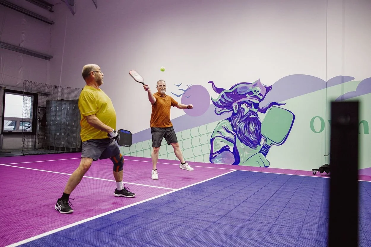 A player mid-air striking the ball on the cushioned VersaCourt surface at Pickleball Cove